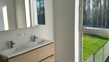 Lichte badkamer met dubbele wastafel, spiegelkast en groot raam met uitzicht op groene tuin en bos.
