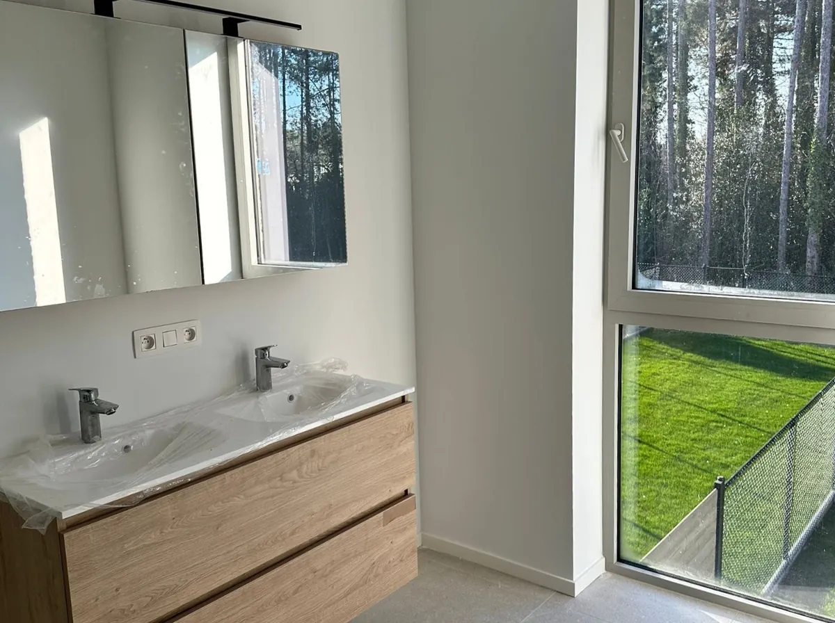 Lichte badkamer met dubbele wastafel, spiegelkast en groot raam met uitzicht op groene tuin en bos.