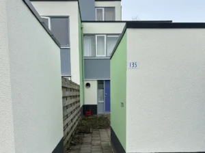 Smal pad tussen twee woningen met frisgroene en witte gevels, strak pleisterwerk. Een rode stoel staat bij een blauwe voordeur.