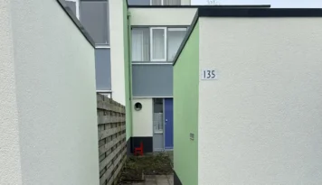 Smal pad tussen twee woningen met frisgroene en witte gevels, strak pleisterwerk. Een rode stoel staat bij een blauwe voordeur.