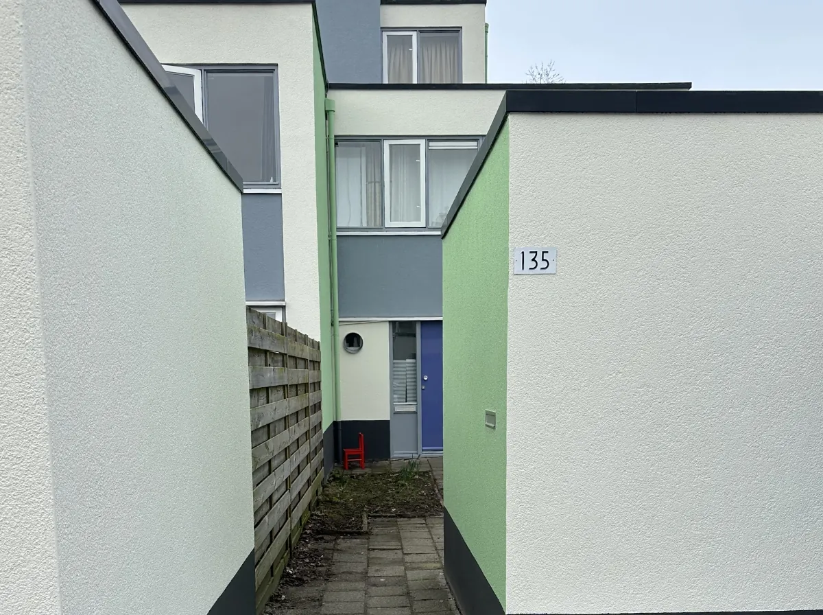 Smal pad tussen twee woningen met frisgroene en witte gevels, strak pleisterwerk. Een rode stoel staat bij een blauwe voordeur.
