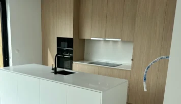 Lichtbruine eikenlook keukenkasten met wit kookeiland en zwarte kraan. De keuken heeft een moderne, strakke uitstraling.