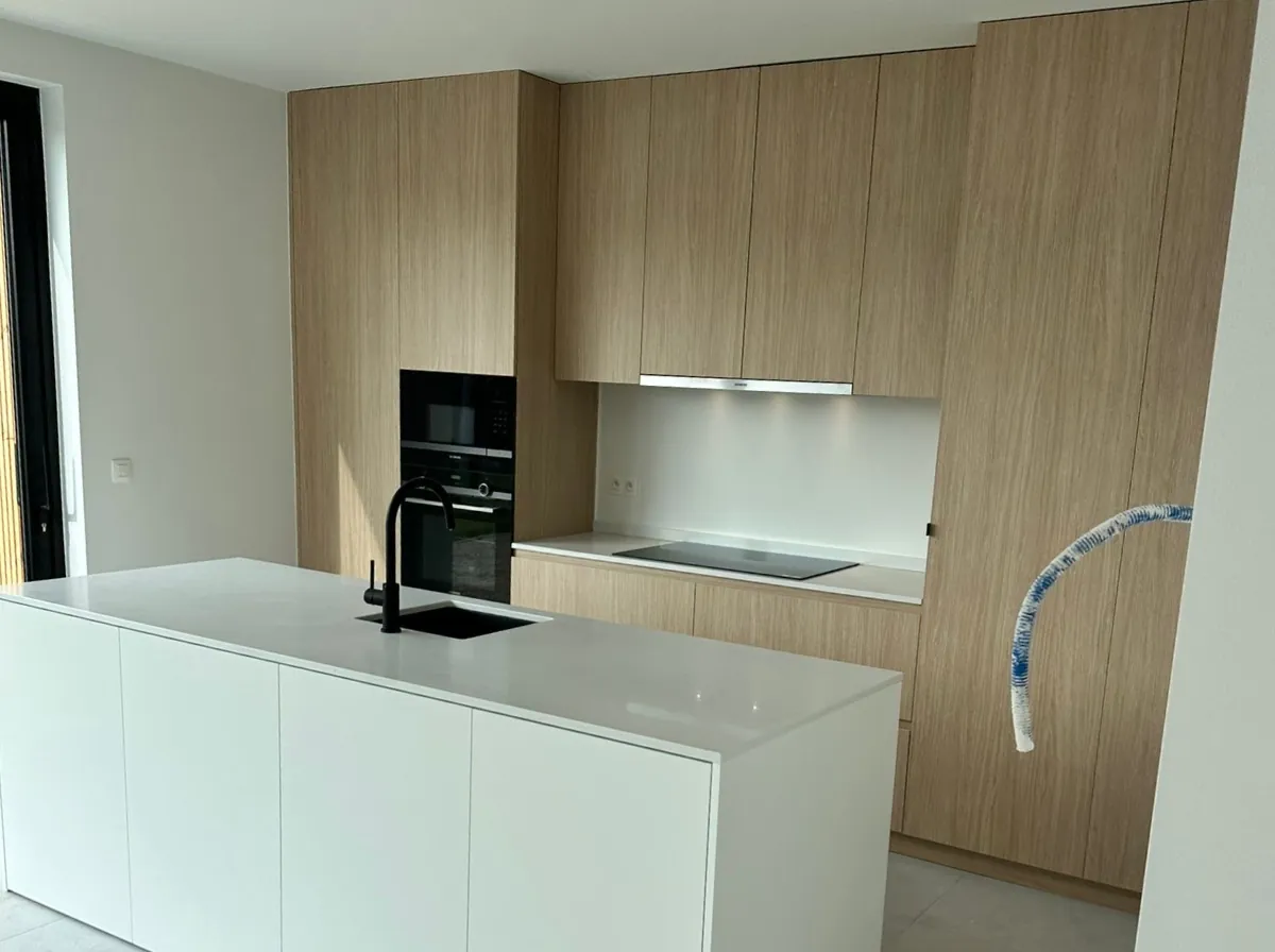 Lichtbruine eikenlook keukenkasten met wit kookeiland en zwarte kraan. De keuken heeft een moderne, strakke uitstraling.